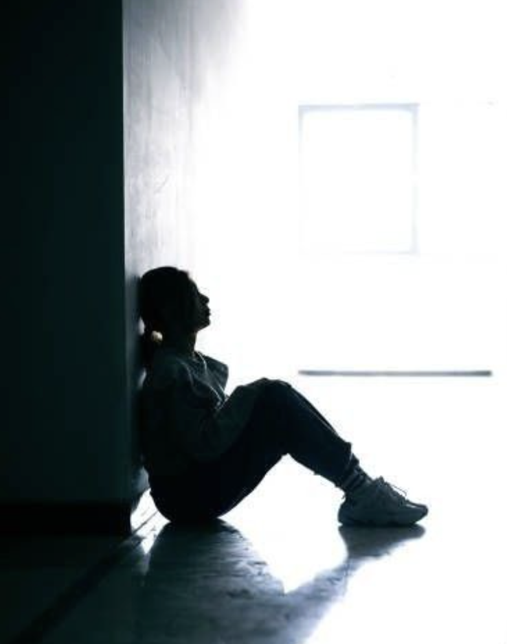 Young person sitting alone against a wall, silhouetted against a distant window